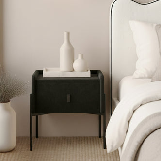 Eliz Velvet Fabric Bedside Table in Black with slender legs, topped with a white tray and ceramic vases, beside a bed with white bedding, exuding a minimalist, contemporary aesthetic.