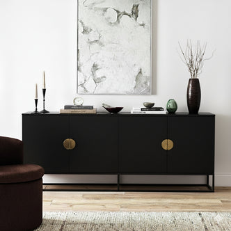 Erick 160cm 4-Door Storage Sideboard Buffet in Black, featuring minimalist brass-tone pulls, is styled with candles, books, and a vase. Positioned on a light-wood floor and beneath abstract art.