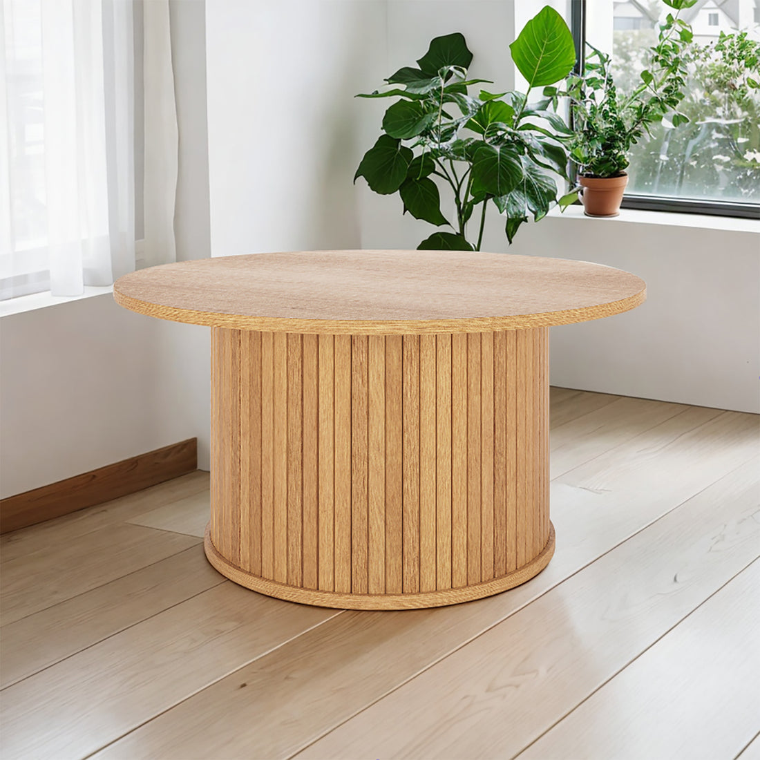 Esmund 90cm Round Coffee Table in Natural, featuring a circular top and fluted base, sits on pale planks near a window, accompanied by houseplants, creating a minimalist and serene interior scene.