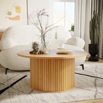 Esmund 90cm Round Coffee Table in Natural features vertical slatted sides, topped with glass jars, pine cones, books, and a ceramic cup, set against a modern living room backdrop.
