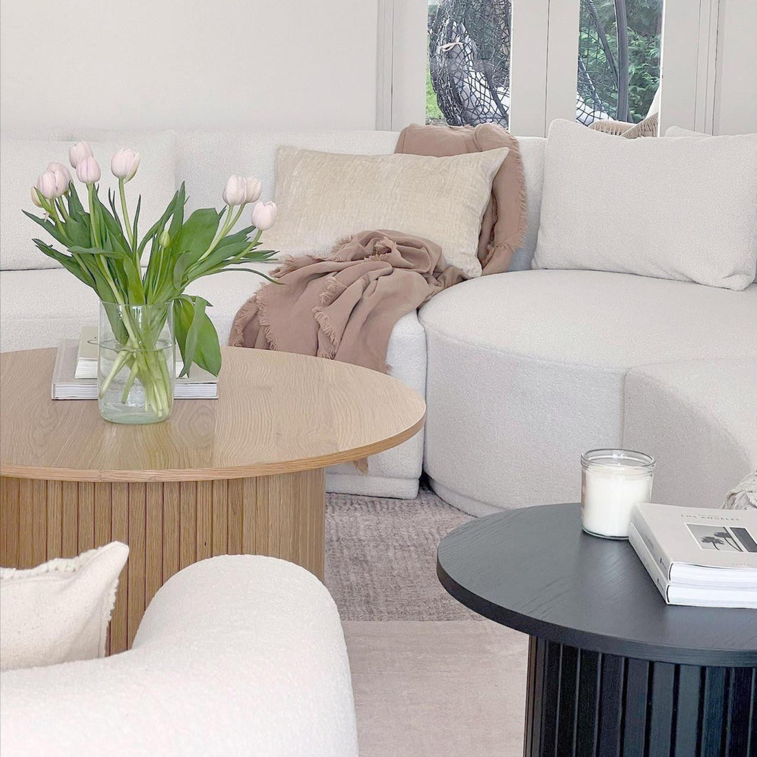 Esmund 90cm Round Coffee Table in Natural, featuring a fluted base, holds a clear vase with pink tulips and stacked books. It's paired with a matching black table in a minimalist living room setting.