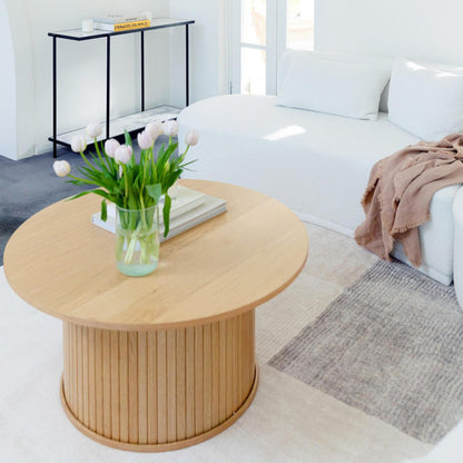 Esmund 90cm Round Coffee Table in Natural features a slatted cylindrical base, topped with books and a glass vase of tulips, harmonizing with a minimalist living area.