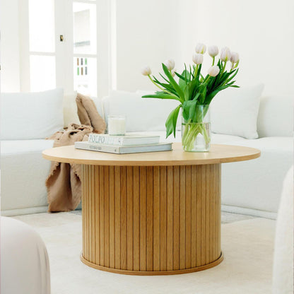 Esmund 90cm Round Coffee Table in Natural features a vertically slatted cylindrical base, topped with two books, a white candle, and a vase of white tulips, creating a minimalist living room vignette.