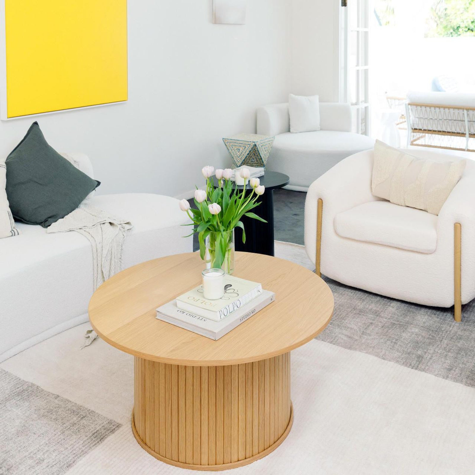 Esmund 90cm Round Coffee Table in Natural, featuring slatted wood design, holds a clear glass vase with tulips, a candle, and design books, central to a minimalist living room setting.