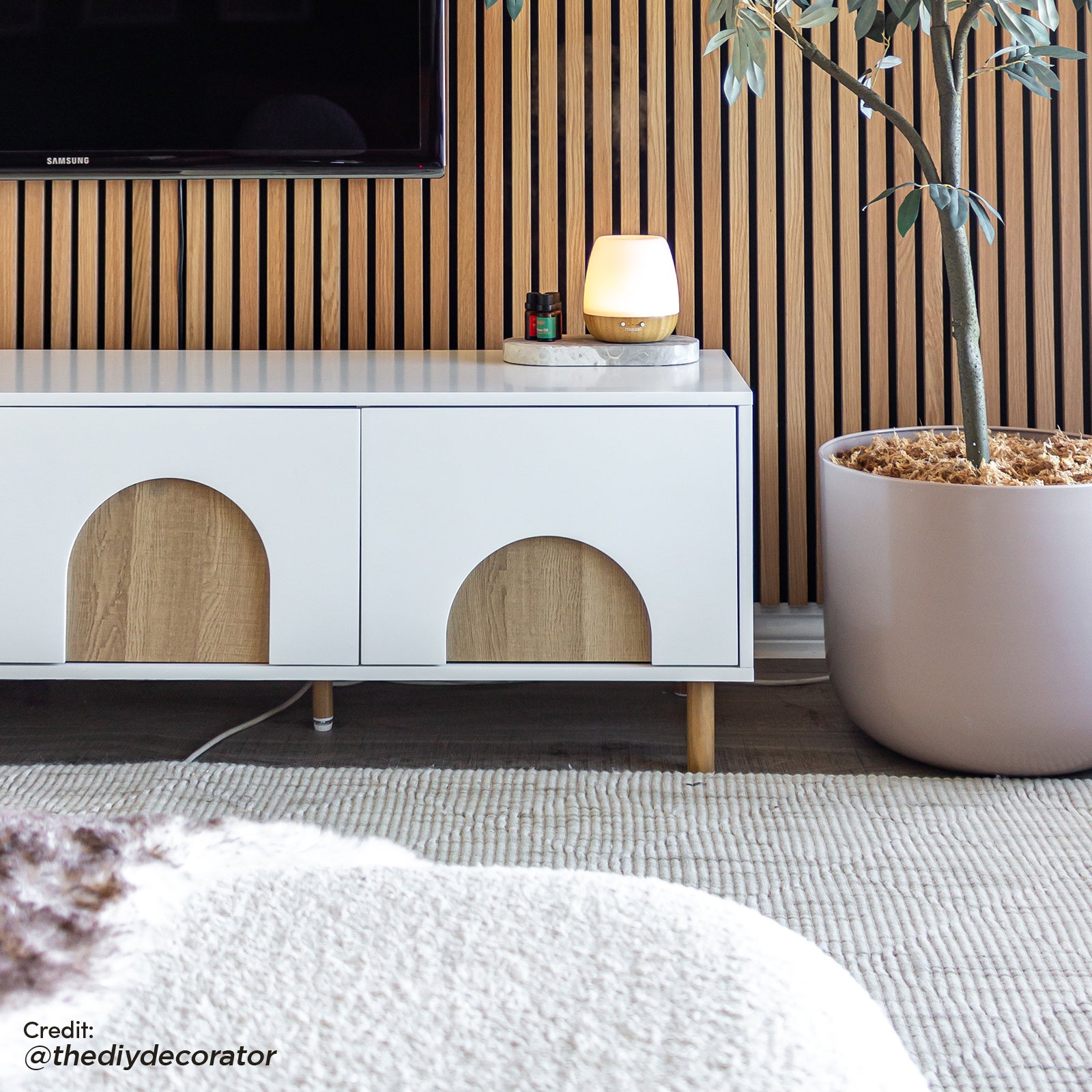 Espur 180cm Entertainment Unit with Drawers in White beneath a wall-mounted TV, featuring half-circle inlays, with essential oils on top, next to a diffuser and large potted tree.