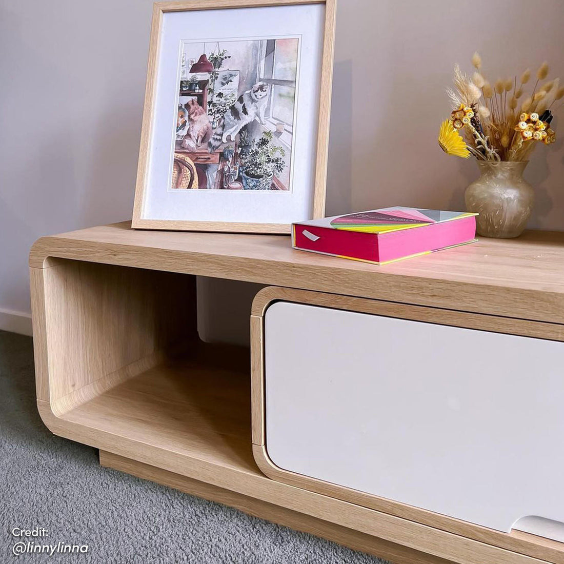 Galya Extendable Storage TV Unit - Oak with open cubby, white sliding panel, adorned with a cat-themed print, pink book, and vase of dried flowers on top.