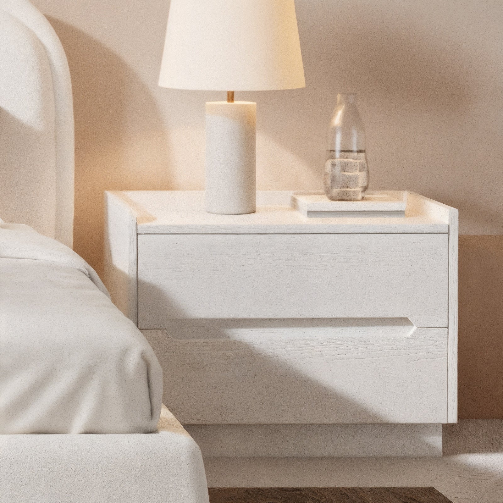 Glorinda 60cm Scratch Resistant Bedside Table in White with two drawers is showcased beside a bed, featuring a light wood finish, a table lamp, books, and a glass carafe.