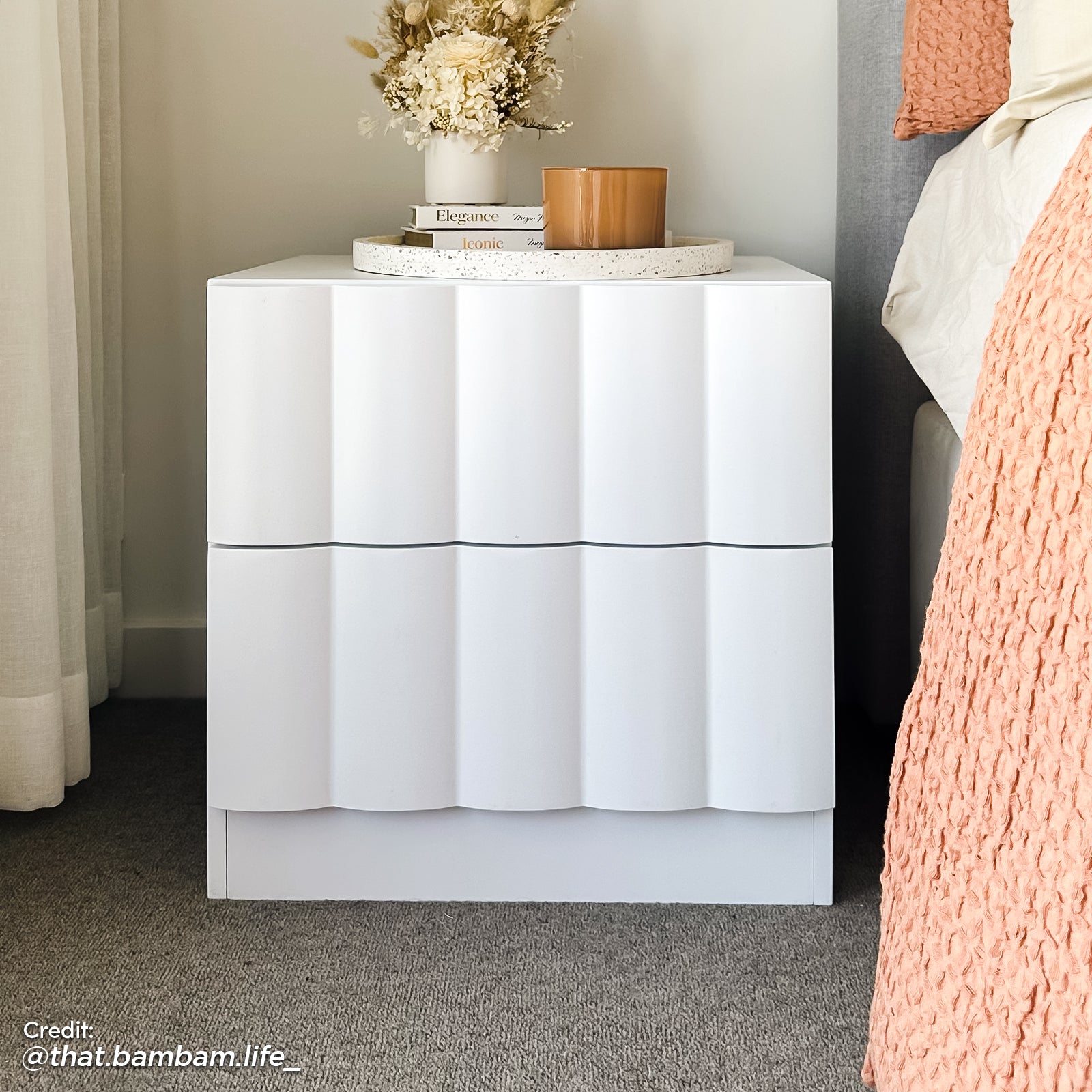 Modern white fluted bedside table with two drawers, styled with a floral arrangement and amber candle in a neutral bedroom setting.