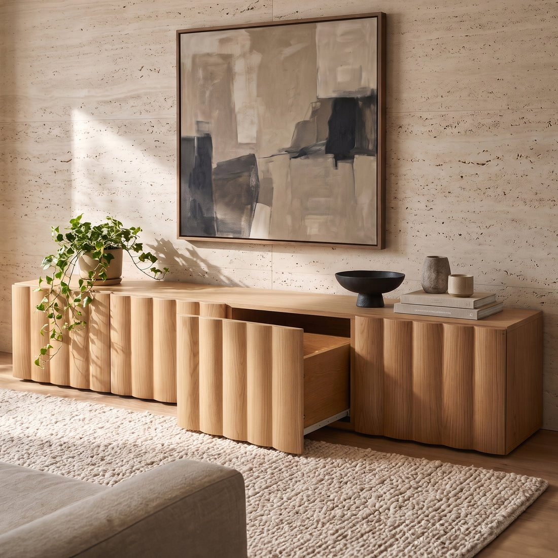 Oak TV unit with vertical fluted front panels and open pull-out drawer, styled with a trailing potted plant, decorative ceramics, and framed abstract wall art against a textured stone wall.