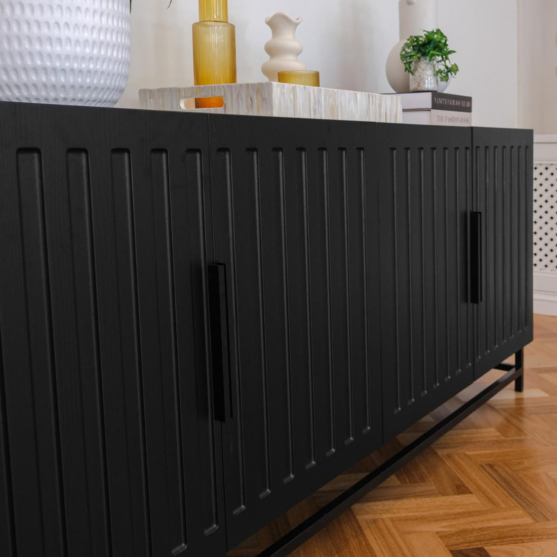 Holly 180cm 4-Door Storage Buffet Sideboard in Black with fluted door detailing and sleek steel legs, adorned with decorative vases and books on a herringbone wood-parquet floor.