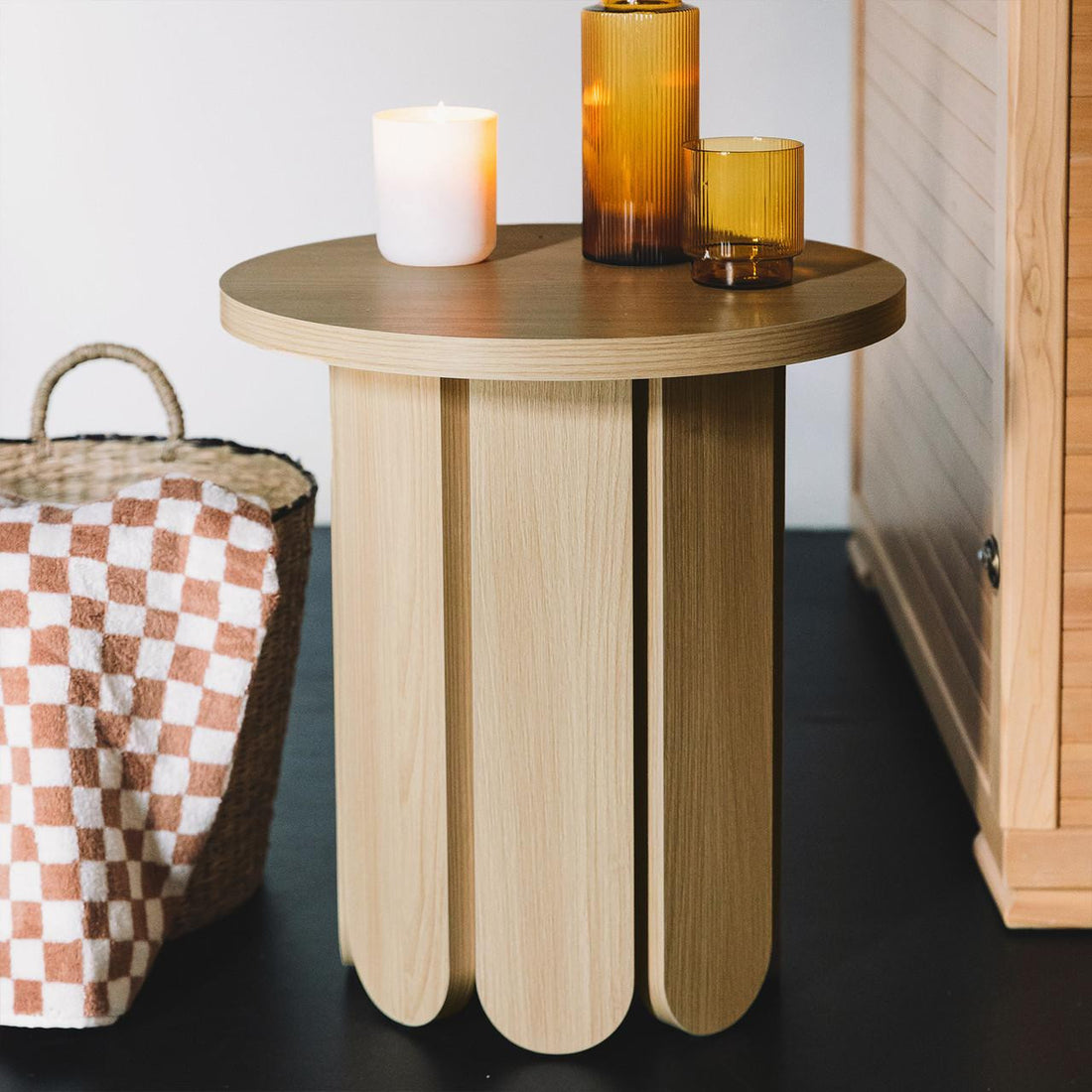 Hugo 45cm Round Side Table with Curve Detail in Natural, featuring a scalloped-edge pedestal base, topped with a lit candle and amber vessels, next to a wicker basket with a blanket.