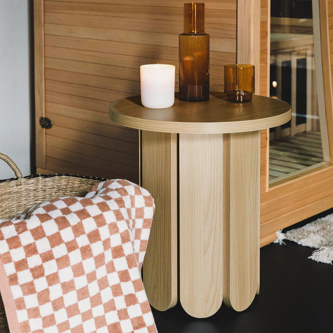 Hugo 45cm Round Side Table with Curve Detail in Natural, featuring light wood vertical slats with scalloped bottoms, adorned with a lit candle, amber vase, and tumbler, creating a cozy, natural setting.