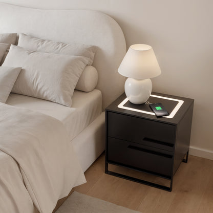 Isha Smart Bedside Table with Wireless Charger in Black features two drawers, a white ceramic lamp, and an integrated wireless charging pad with a smartphone charging, enhancing modern bedroom convenience.