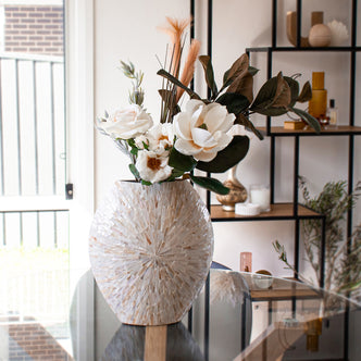 Laila 36cm Mother of Pearl Inlay Vase - Cream Beige on a glass table with white blooms, green foliage, and dried grasses, set against a shelf with decor items and a sunlit background.