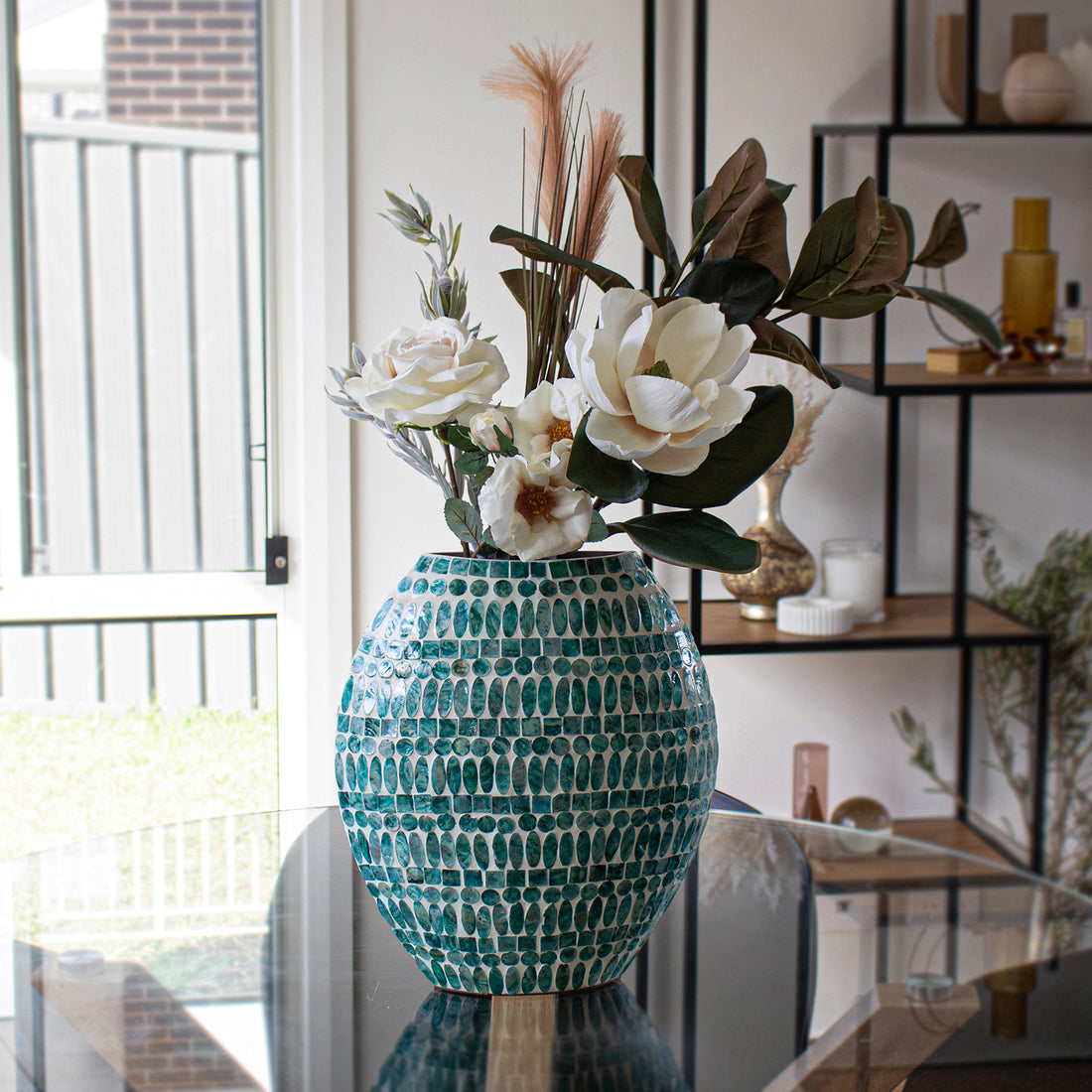 Laila 36cm Mother of Pearl Inlay Vase - Jade Green, features a mosaic design with turquoise oval tiles, filled with faux white blooms and greenery, positioned on a glass table in a bright room.
