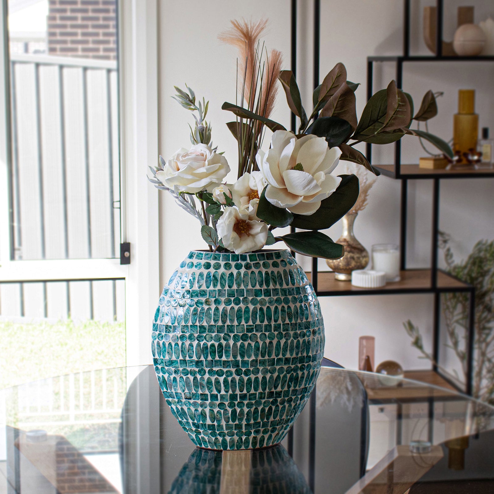 Laila 36cm Mother of Pearl Inlay Vase - Jade Green, features a mosaic design with turquoise oval tiles, filled with faux white blooms and greenery, positioned on a glass table in a bright room.