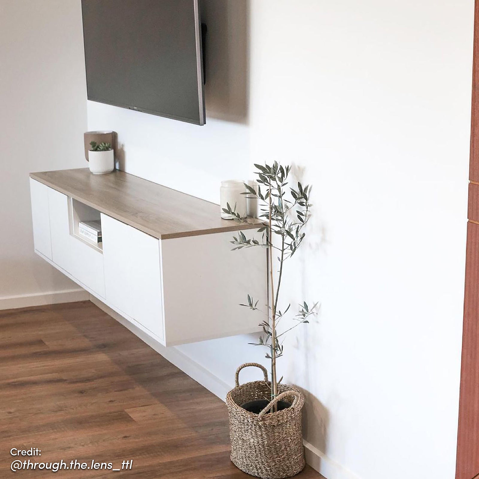 Leslie 180cm Floating Entertainment Unit in White & Natural beneath a wall-mounted TV, featuring sleek white doors, light wood-grain top, and minimalist decor—small planter, ceramic jar, and woven basket with a plant.