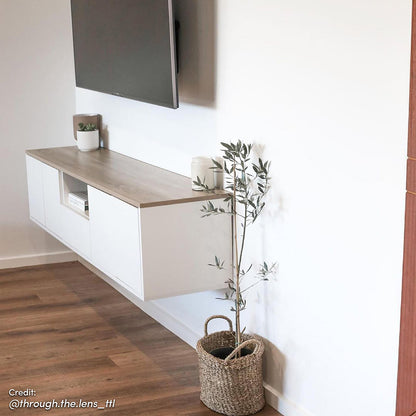 Leslie 180cm Floating Entertainment Unit in White & Natural beneath a wall-mounted TV, featuring sleek white doors, light wood-grain top, and minimalist decor—small planter, ceramic jar, and woven basket with a plant.