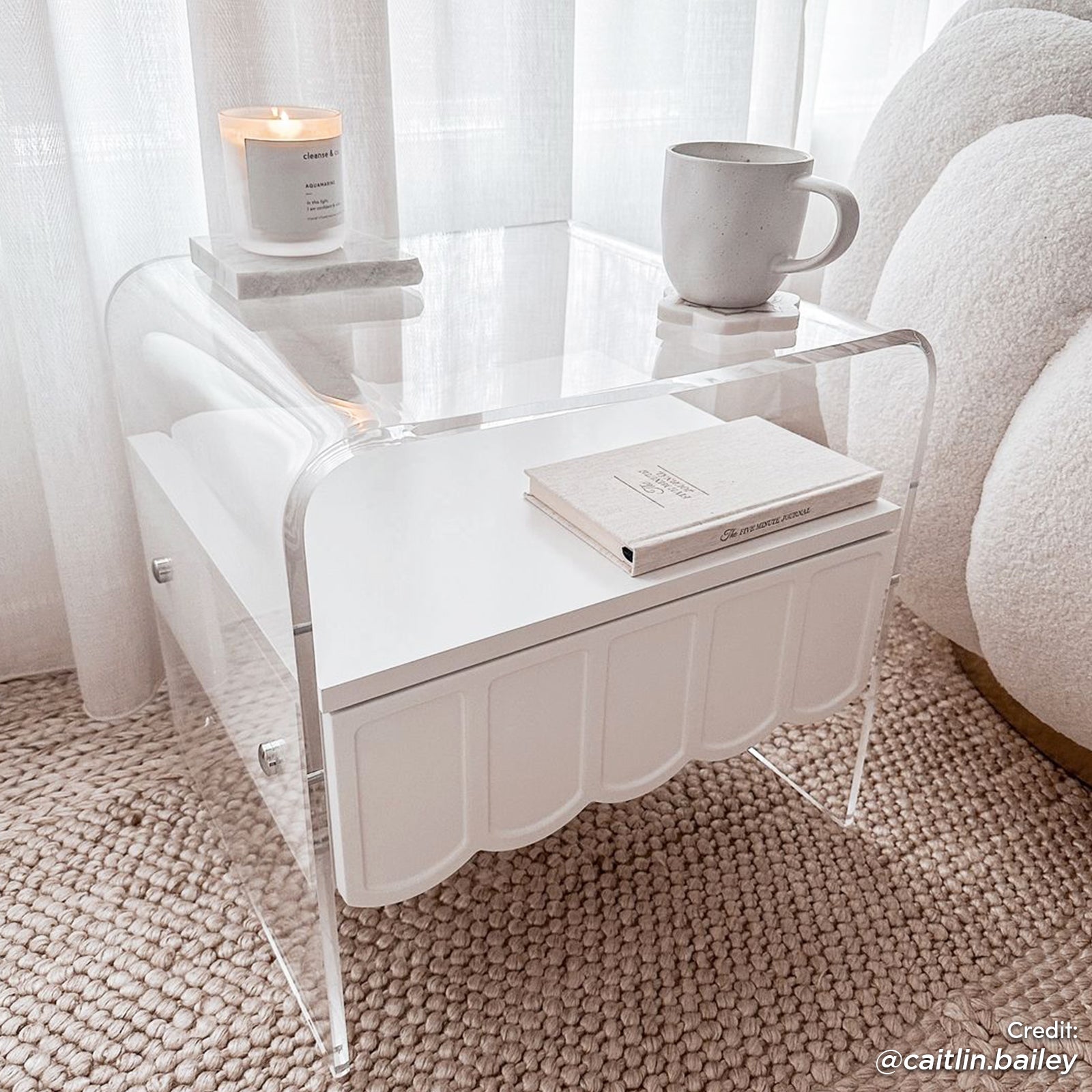 Magdalena 45cm Scalloped Acrylic Bedside Table in White, featuring a clear U-shaped acrylic frame, scalloped-edge removable drawer, on a natural fiber rug, topped with a candle, mug, and journal.