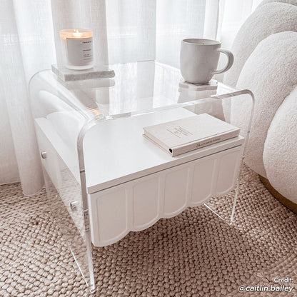 Magdalena 45cm Scalloped Acrylic Bedside Table in White, featuring a clear U-shaped acrylic frame, scalloped-edge removable drawer, on a natural fiber rug, topped with a candle, mug, and journal.