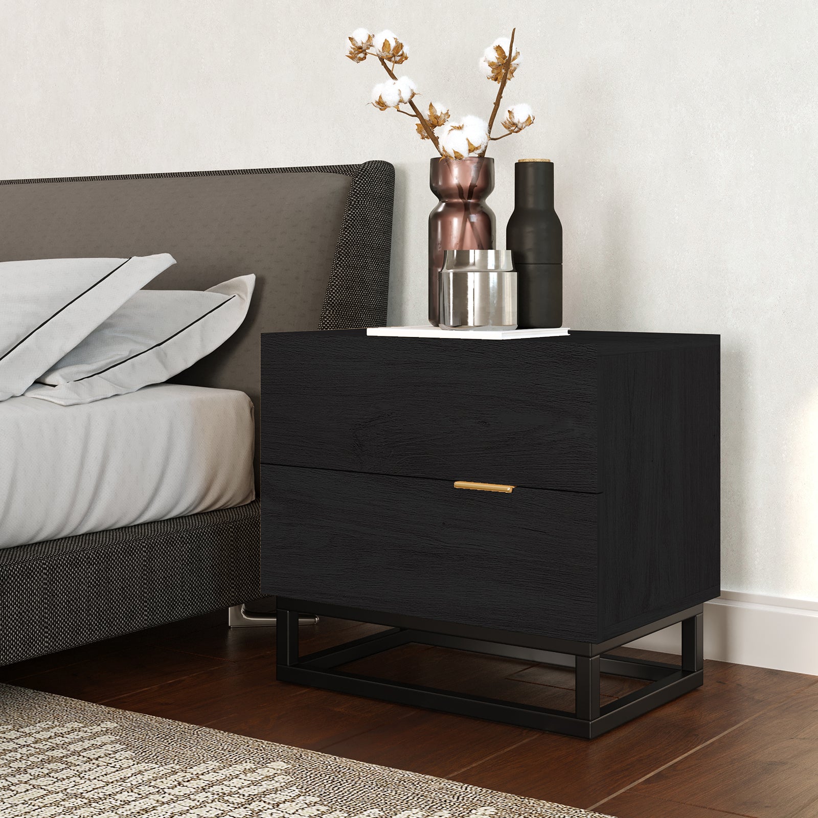 Manuel 2 Drawer Storage Bedside Table Black with gold handles, featuring two drawers on a black metal base, adorned with decorative vessels on top, beside an upholstered bed in a modern bedroom setting.