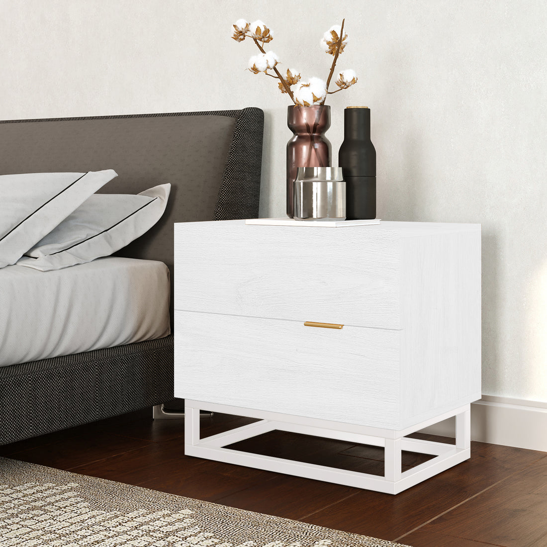 Manuel 2 Drawer Storage Bedside Table White beside a bed, featuring a minimal metal base, gold handles, and topped with decorative vessels, set on warm wood flooring with a patterned rug.