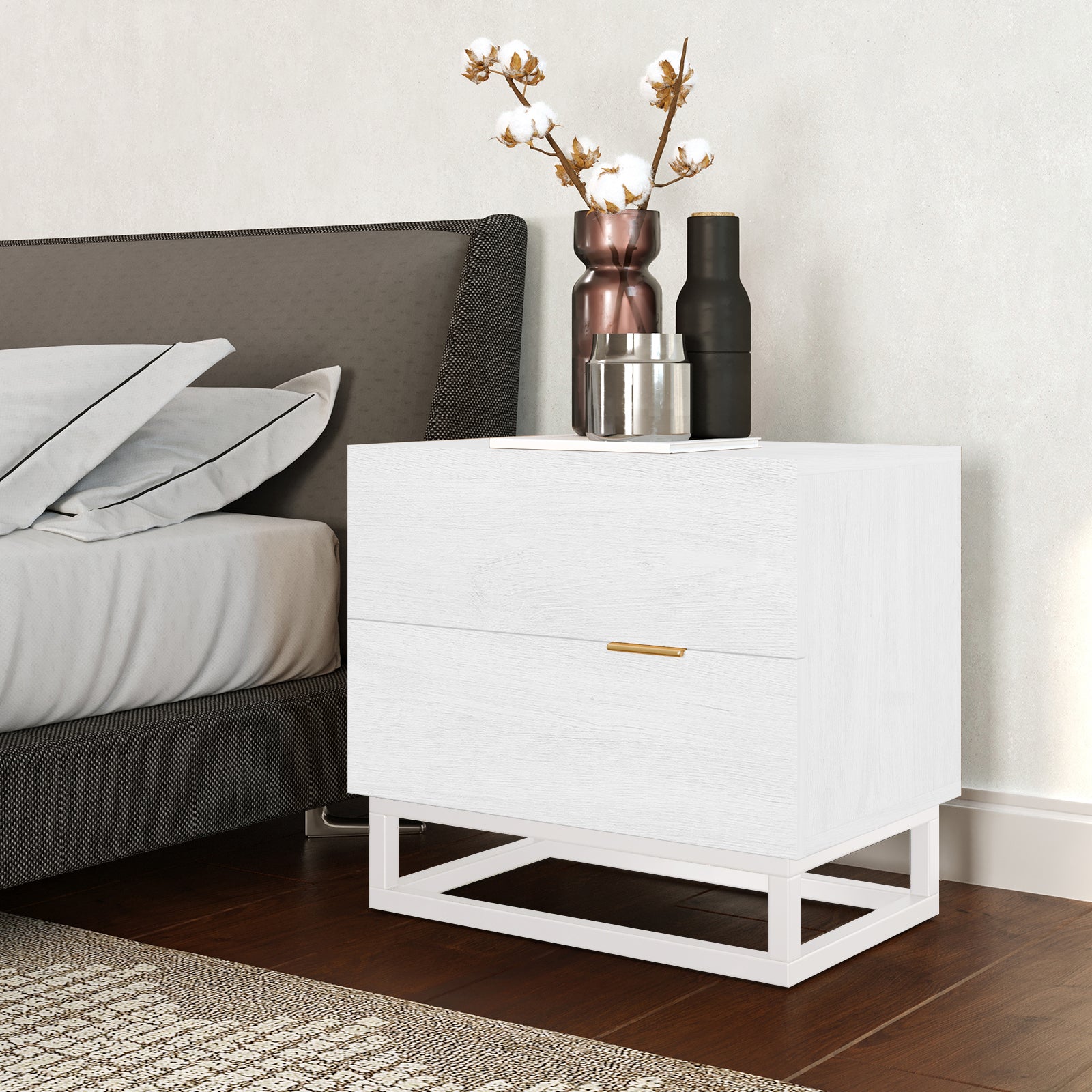 Manuel 2 Drawer Storage Bedside Table White beside a bed, featuring a minimal metal base, gold handles, and topped with decorative vessels, set on warm wood flooring with a patterned rug.