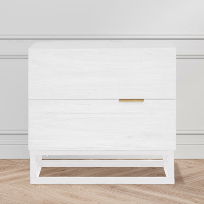 Manuel 2 Drawer Storage Bedside Table White with white wood grain finish, two drawers, slim brass-tone handle, and a simple white metal frame, against a light-grey wall on oak-tone floor.