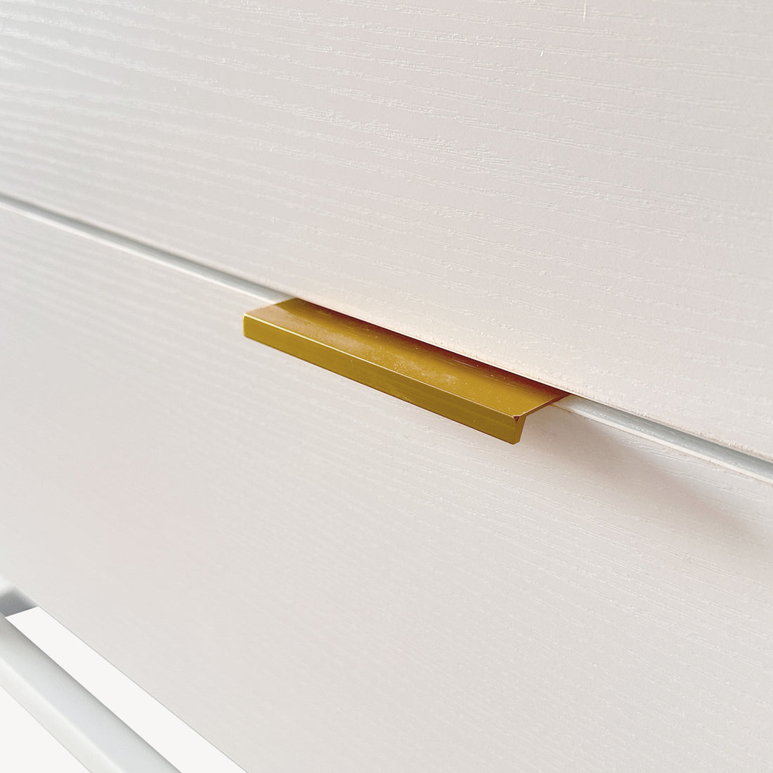Close-up of the Manuel 2 Drawer Storage Bedside Table White, featuring a white wood-grain-textured drawer front with a slim rectangular gold metal pull handle near the top edge.