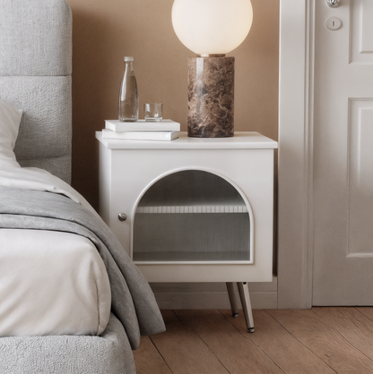 Matteau 46cm Bedside Table with Glass Door in White, featuring a sleek design with metal legs and an elegant fluted glass door, stands beside a bed in a modern bedroom setting.
