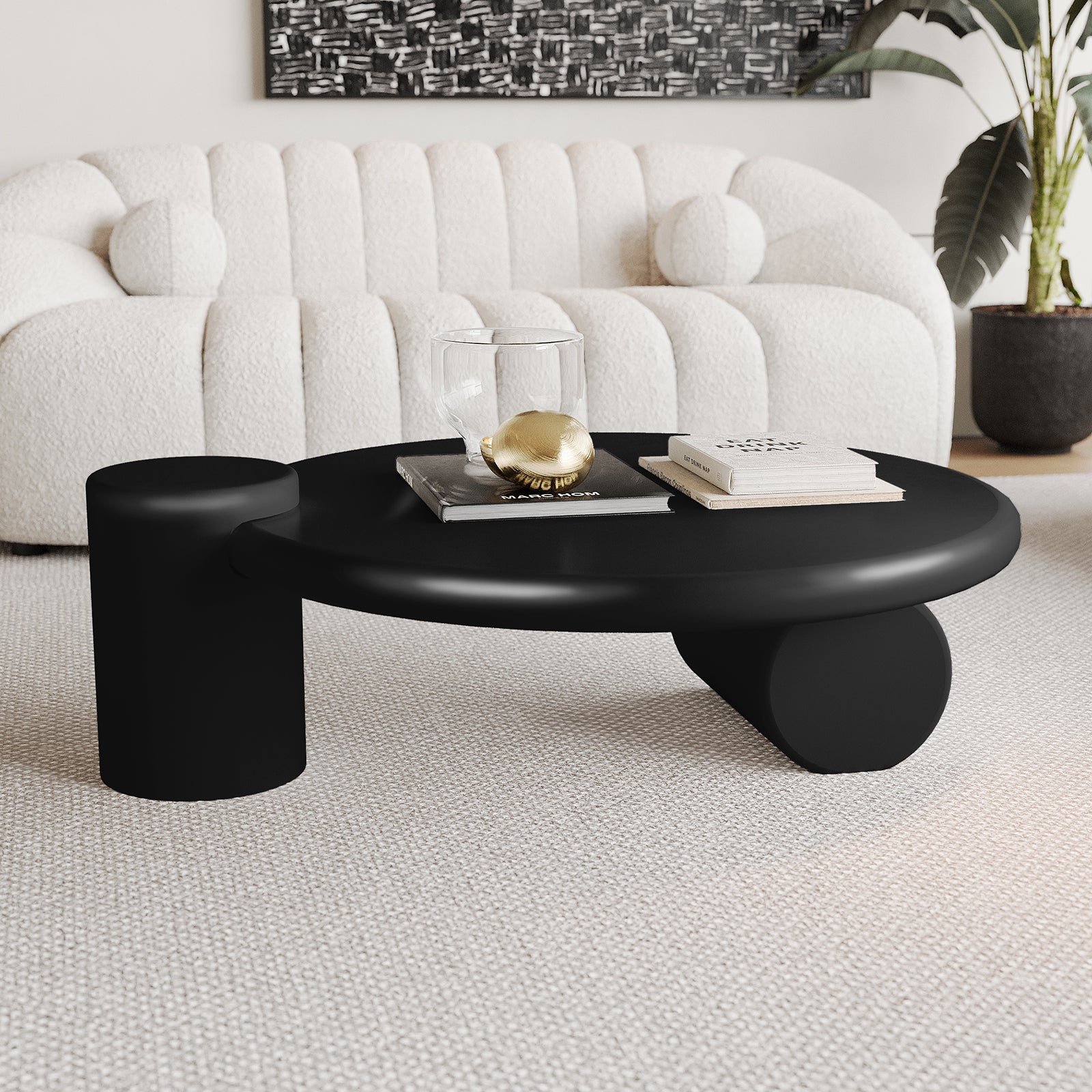 Nando 100cm Modern Round Coffee Table in Black featuring dual cylindrical supports, a glass tumbler, gold decorative orb, and books. Complements an off-white sofa and abstract artwork in a minimalist setting.