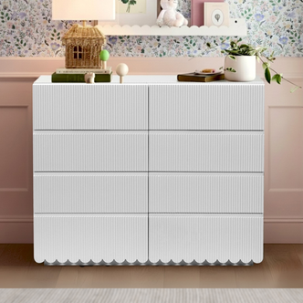 Nasrin 120cm Lowboy Dresser with Drawers in White, featuring six drawers and a scalloped base, sits against a floral wall. Decor includes a wicker lamp, books, spheres, plant, and shelf items.