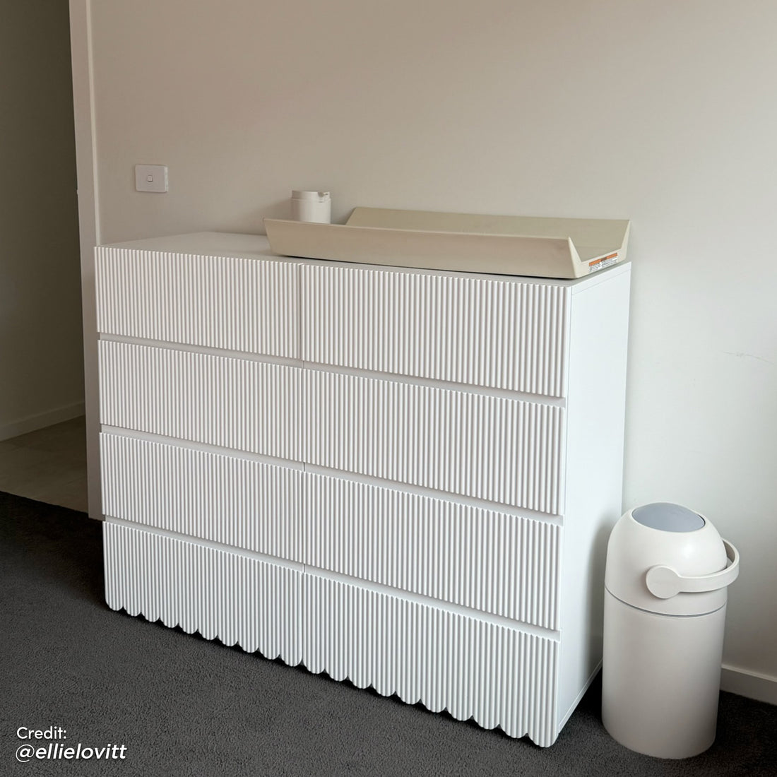 Nasrin 120cm Lowboy Dresser with Drawers in White features a minimalist design with vertically ribbed drawer fronts and scalloped base, topped with a beige changing pad, next to a white diaper pail.