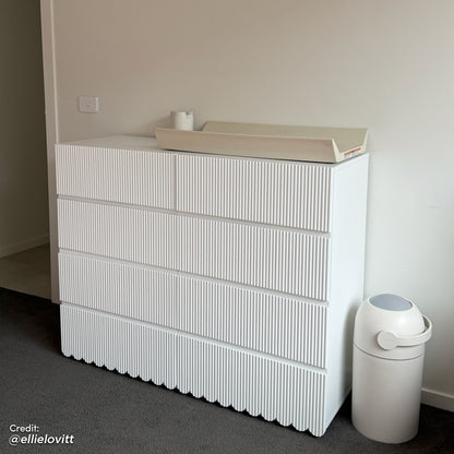 Nasrin 120cm Lowboy Dresser with Drawers in White features a minimalist design with vertically ribbed drawer fronts and scalloped base, topped with a beige changing pad, next to a white diaper pail.