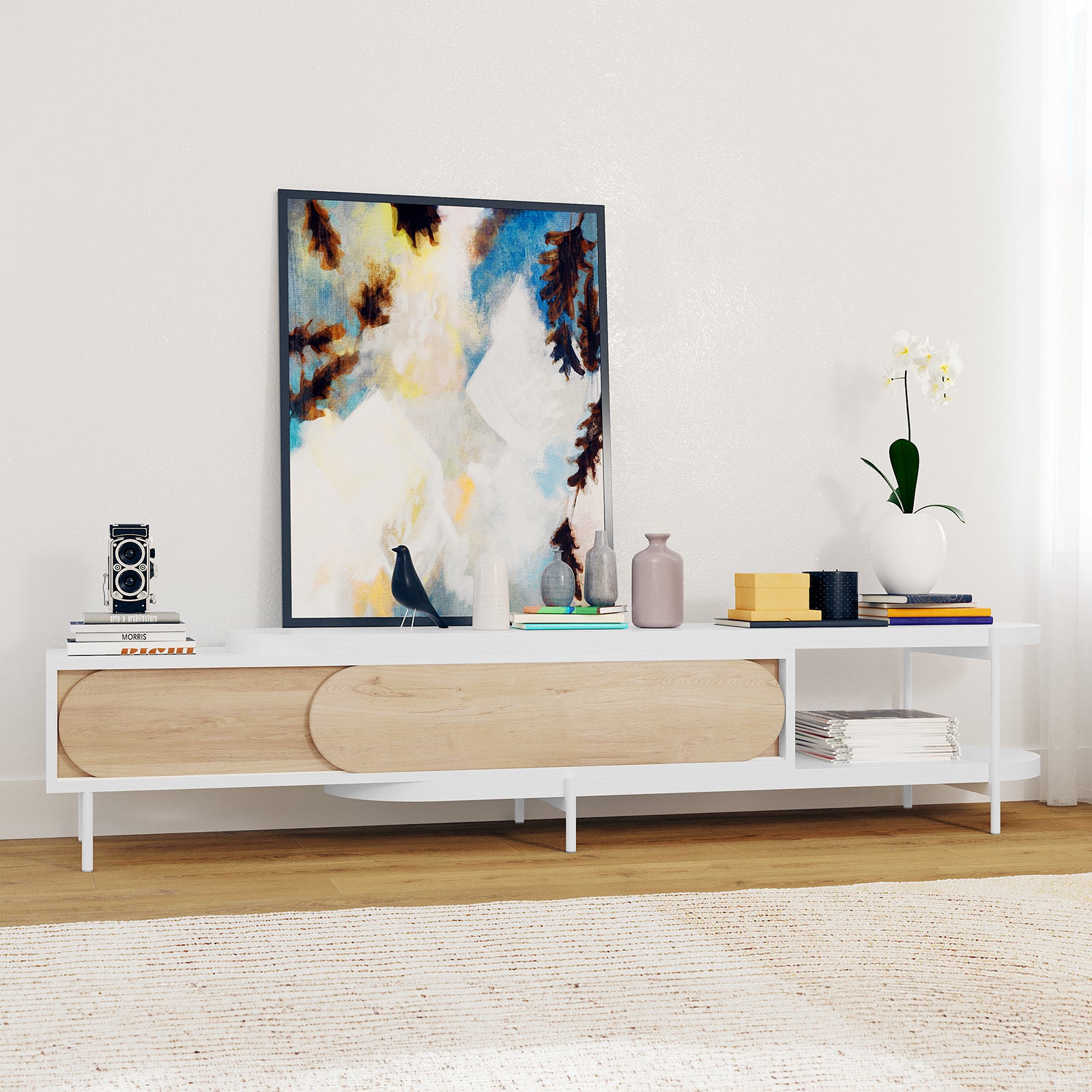 Novia 220-270cm Extendable Entertainment TV Unit in White, featuring sliding oval-fronted wooden doors, open cubby with magazines, slender white legs, and a chic top adorned with books, art, and decor.