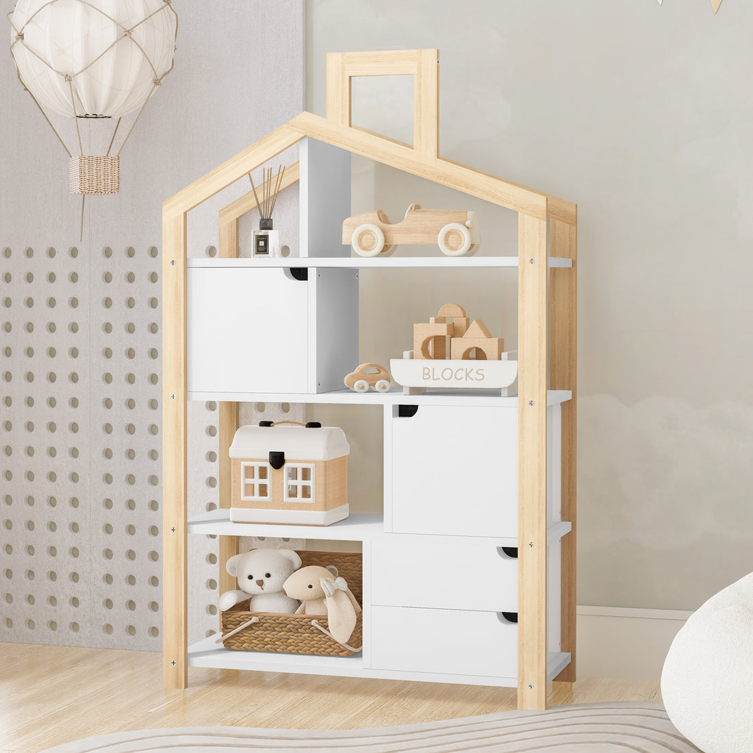 Quinta Shelf Unit with Storage White & Natural, featuring a house-shaped design with toys and blocks displayed on its shelves, a woven basket with plush toys, and a decorative hot-air-balloon mobile above.