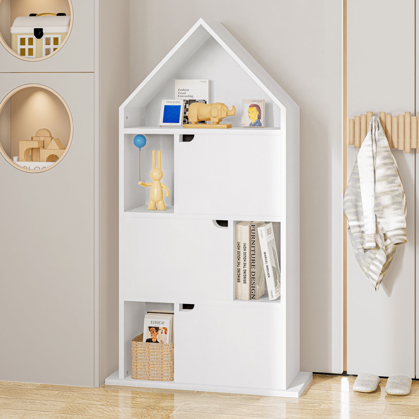 Quinta Bookshelf with Cabinet White shaped like a house, featuring six cubbies with books, toys, and a sculpture, alongside a jacket on a wall-mounted rack and slippers below.