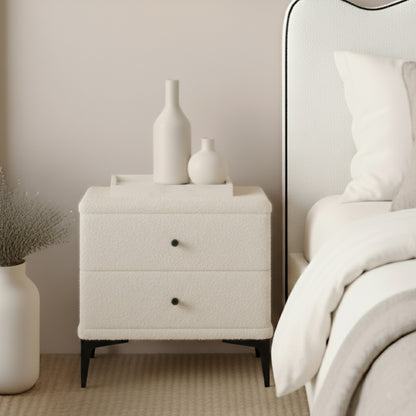 Shona Upholstered Boucle Bedside Table in Cream White with two drawers, slender black metal legs, and a minimalist tray holding ceramic vessels, set beside a bed with white linens.