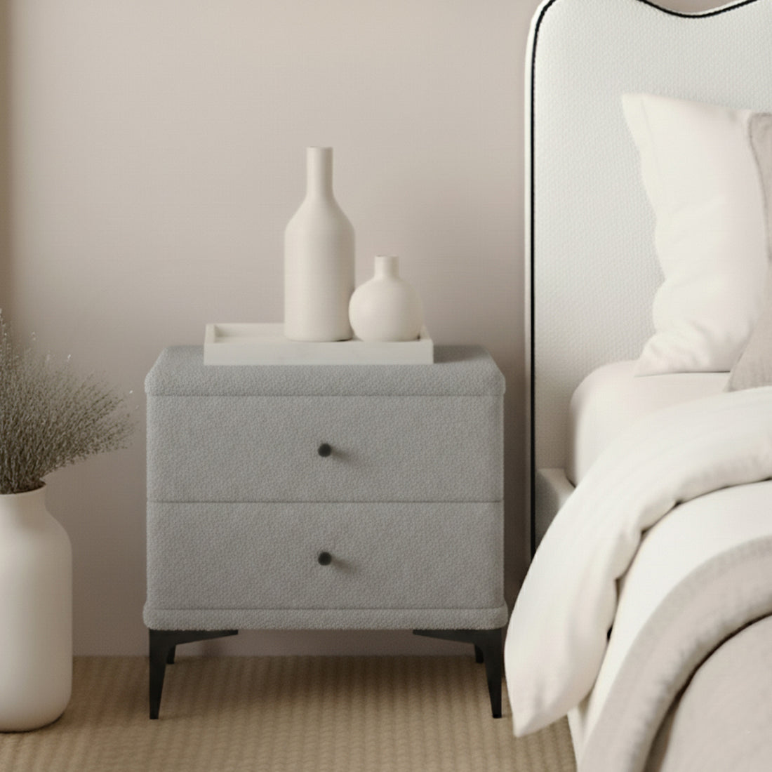 Shona Boucle Upholstered Bedside Table in Grey beside a bed, featuring two drawers with black knobs, slender black legs, and topped with a white tray and ceramic vases for a minimalist look.