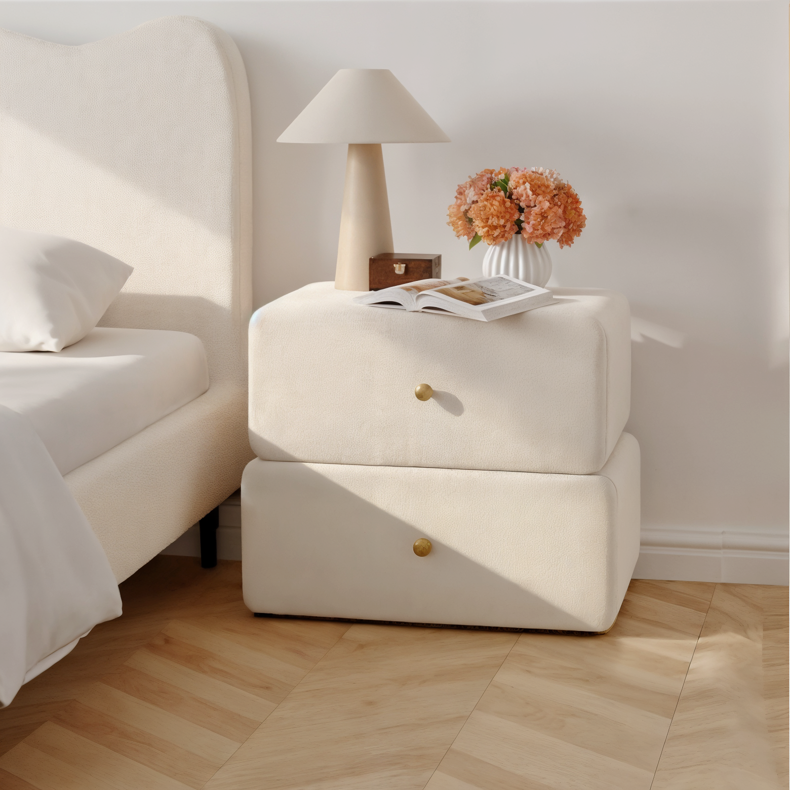 Tyla Velvet Bedside Table with Drawers in Beige, featuring a cream-upholstered finish, brass knobs, and topped with a beige lamp, wooden box, open book, and a vase of orange-peach flowers.