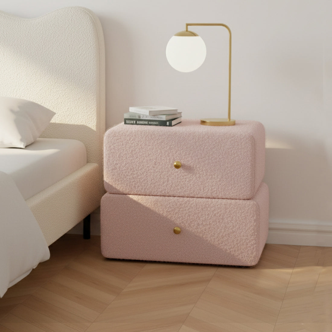 Tyla Boucle Upholstered Bedside Table in Light Pink, featuring rounded edges, brass knobs, and dual drawers, topped with a brass lamp and books, set on herringbone wood flooring.