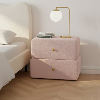 Tyla Boucle Upholstered Bedside Table in Light Pink, featuring rounded edges, brass knobs, and dual drawers, topped with a brass lamp and books, set on herringbone wood flooring.