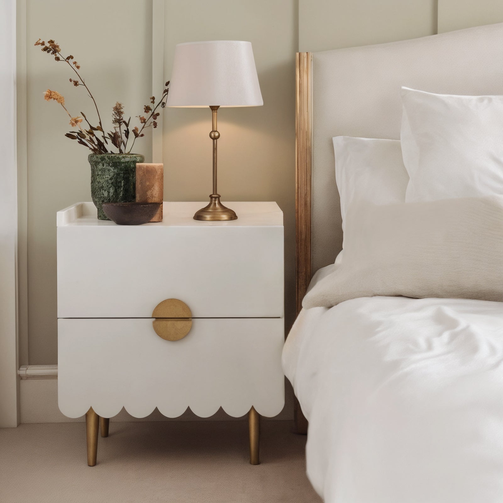Venroy 45cm Bedside Table with 2 Drawers in White, featuring scalloped trim, brass-tone hardware and legs, topped with a brass lamp, green vase with dried stems, candle, and bowl.