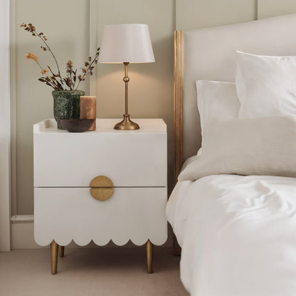 Venroy 45cm Bedside Table with 2 Drawers in White, featuring scalloped trim, brass-tone hardware and legs, topped with a brass lamp, green vase with dried stems, candle, and bowl.