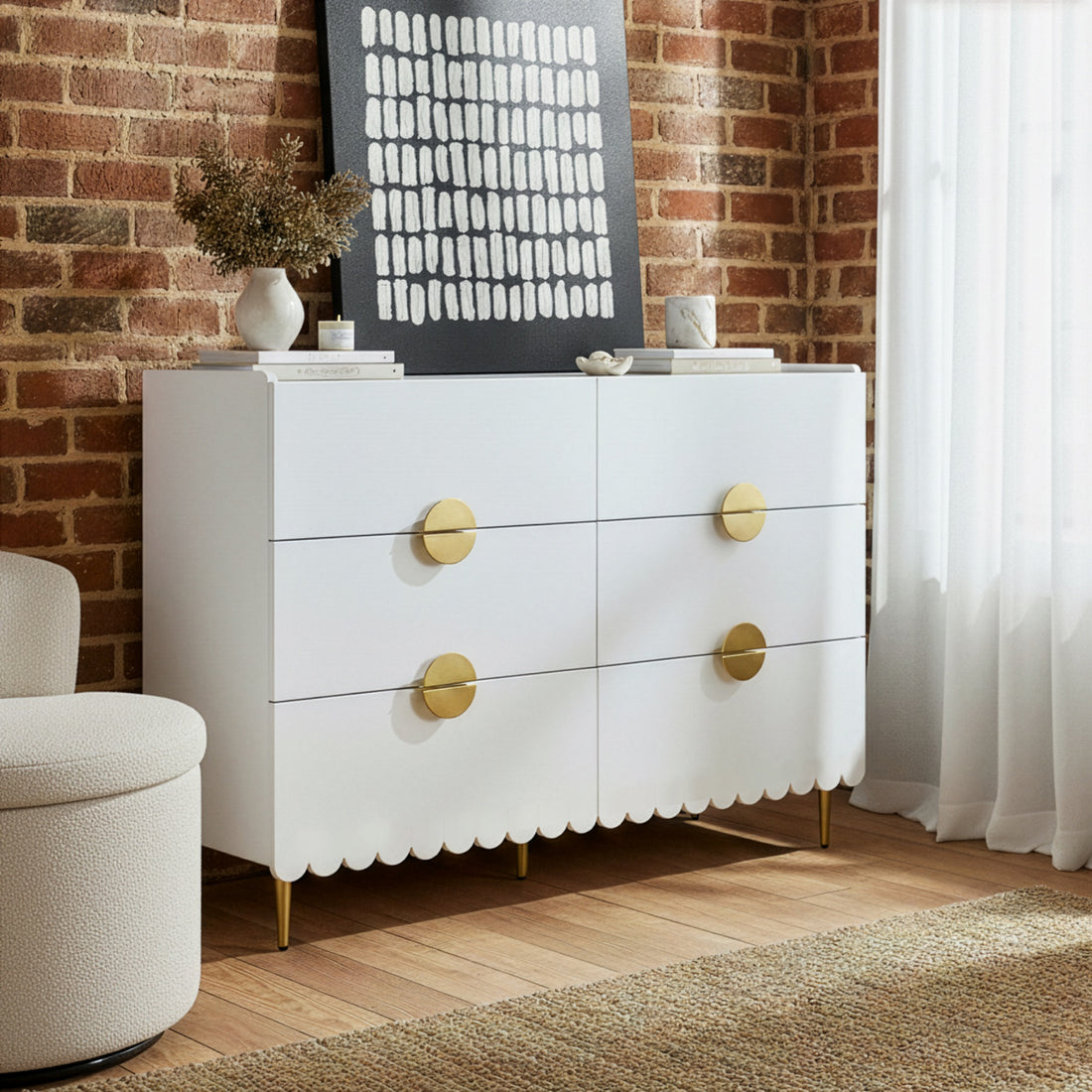 Venroy 120cm Lowboy with 8 Drawers in White features a scalloped apron, brass legs, circular pulls, topped with art and decor, set against an exposed brick wall.