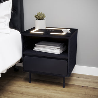 Zayn Smart Bedside with Wireless Charger in Black featuring a potted succulent, smartphone on LED-lit wireless pad, open shelf with books, and a closed drawer, all on slim metal legs.