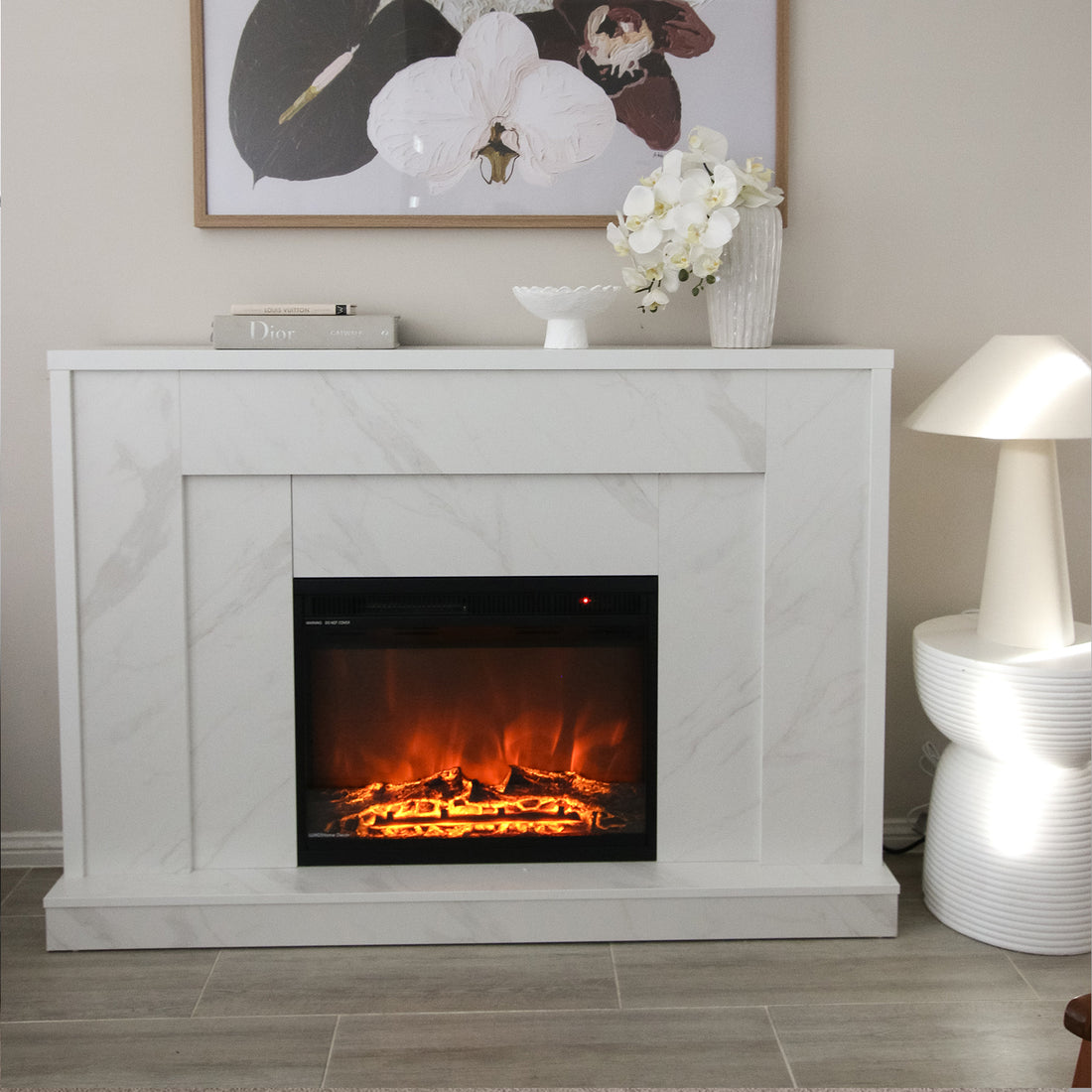 Armidale 132cm 1600W LED Electric Fireplace in White Marble set in a mantel with glowing logs and flickering flames, topped with books, a vase with orchids, and a botanical print above.