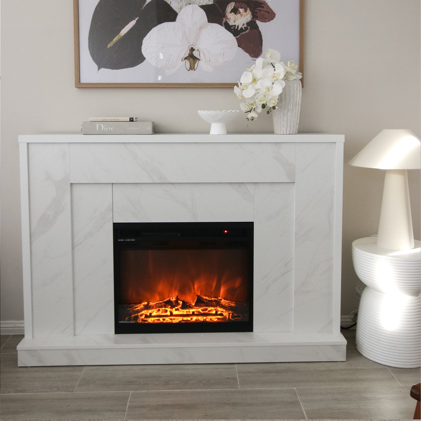 Armidale 132cm 1600W LED Electric Fireplace in White Marble set in a mantel with glowing logs and flickering flames, topped with books, a vase with orchids, and a botanical print above.
