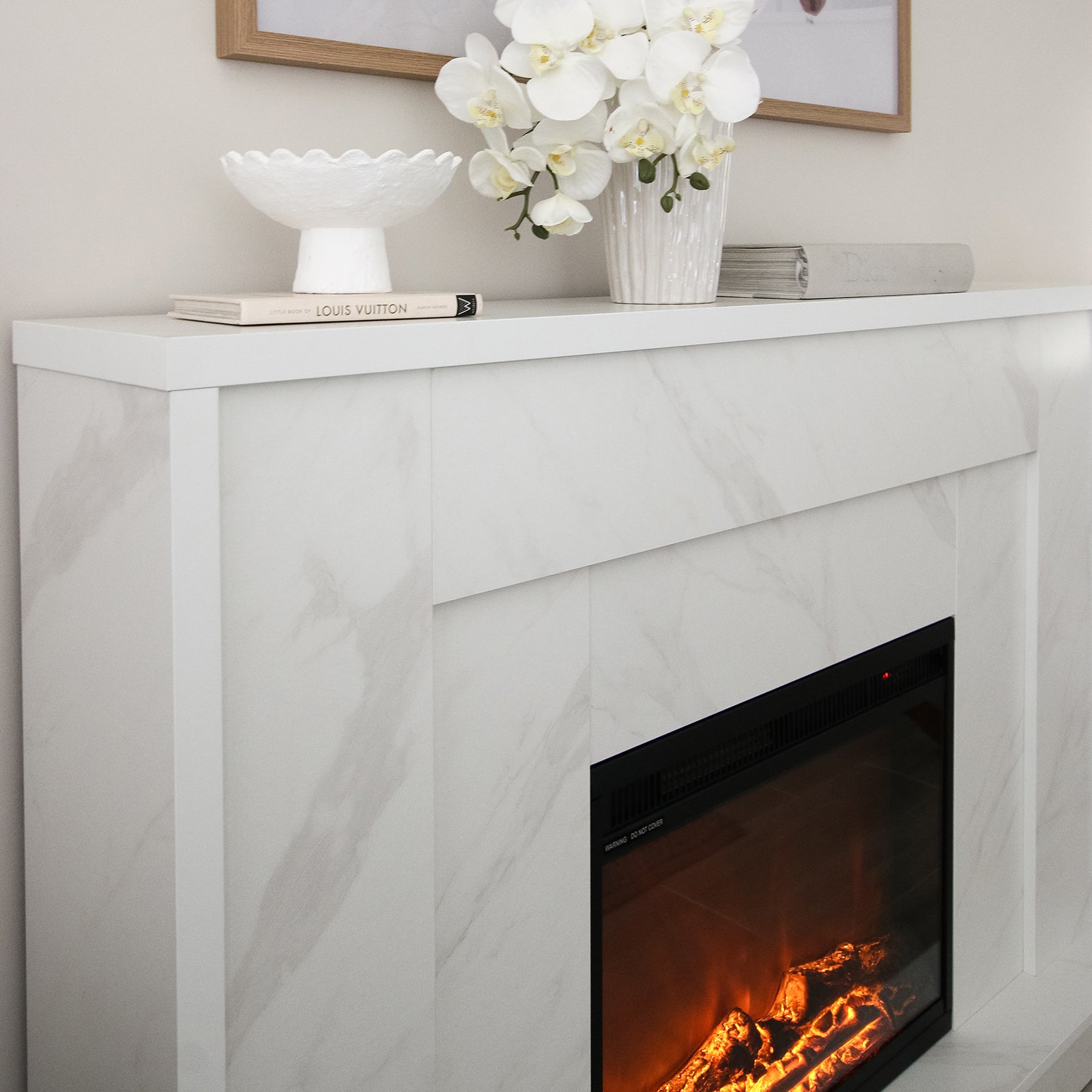 Armidale 132cm 1600W LED Electric Fireplace in White Marble, featuring a light mantel adorned with a decorative bowl, books, orchid, and magazine; glowing faux-log embers providing warm ambiance.