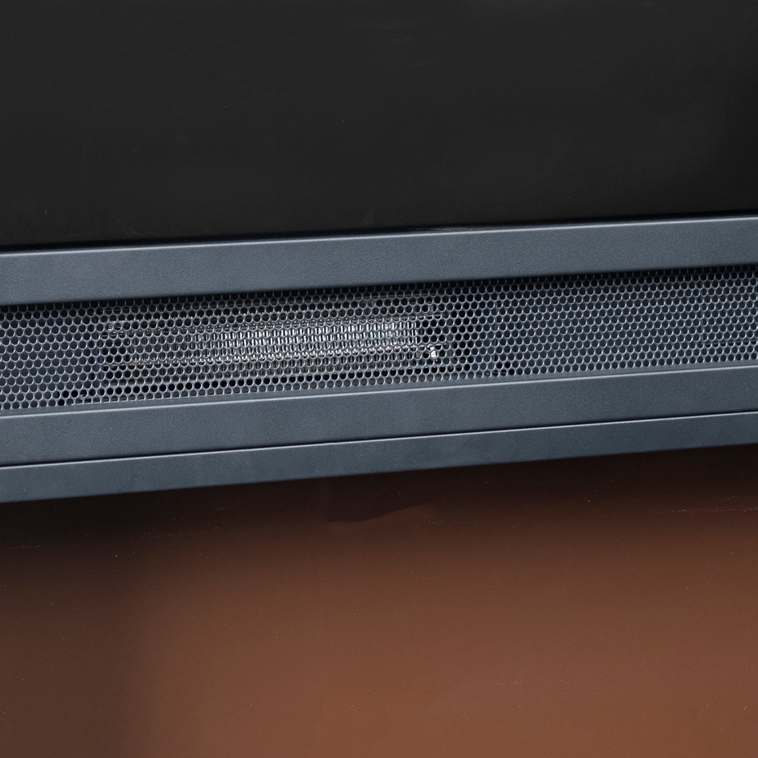 Close-up of Oprah 1950W Free Standing Electric Mantel Fireplace in Black, showing black bezel and perforated metal grille, likely part of the speaker or vent, on a wooden surface.